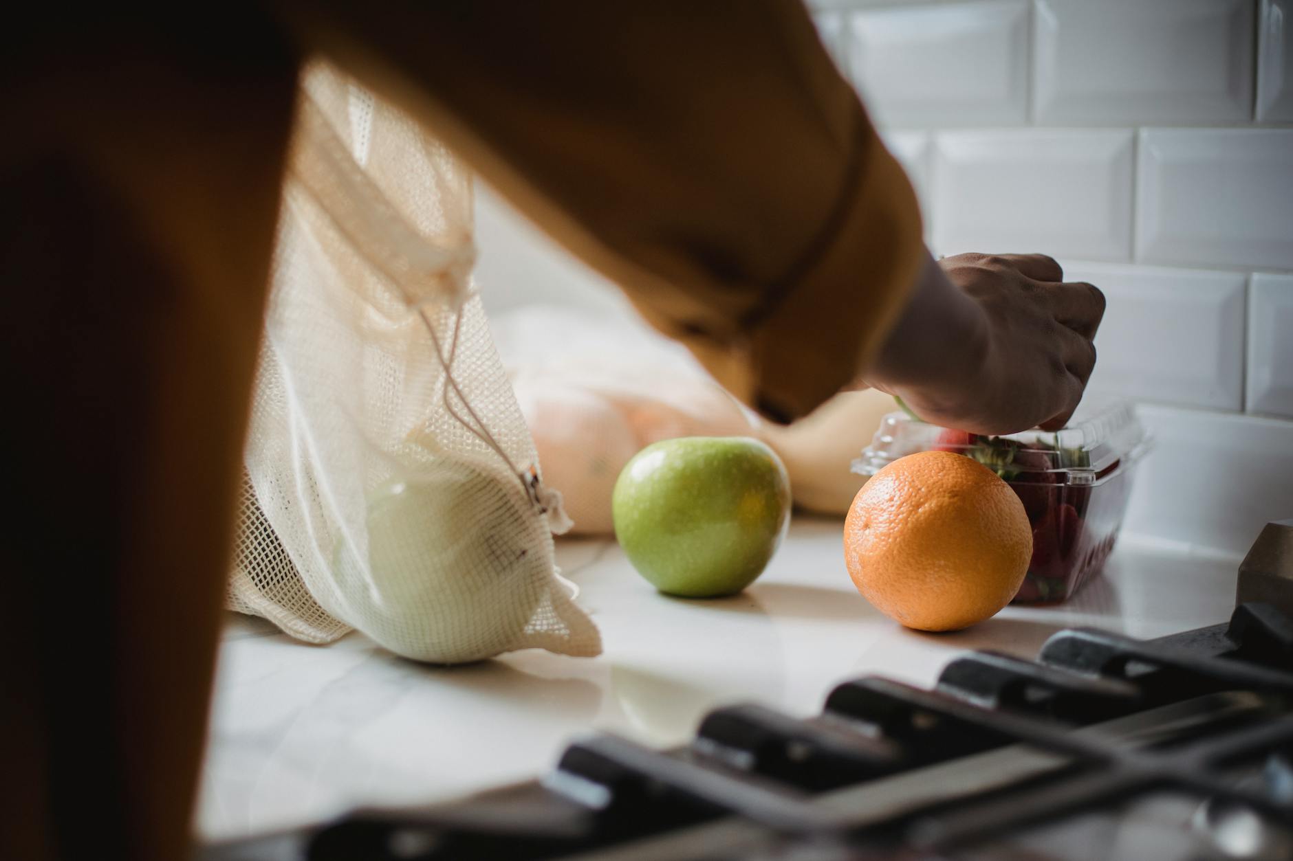 Person unpacking eco-friendly bag with fresh fruits in a modern kitchen. (Photo by Ketut Subiyanto on Pexels)