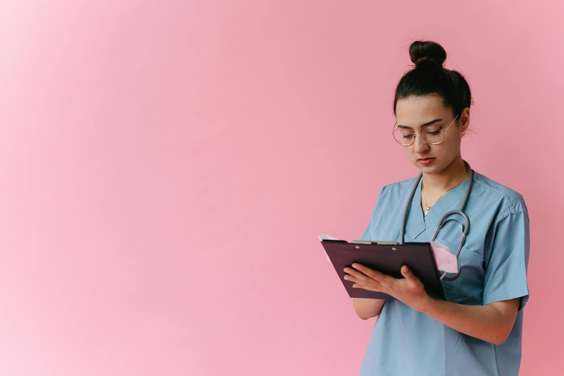 Professional nurse reviewing patient notes on clipboard with pink background, in studio setup. (Photo by Thirdman on Pexels)