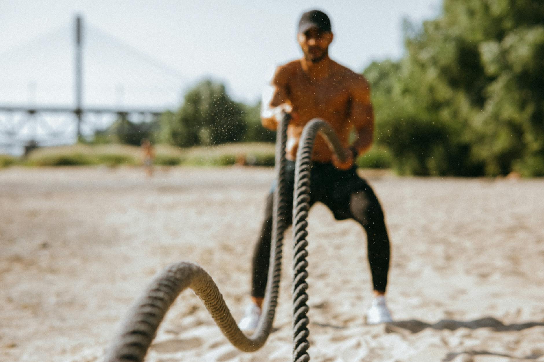 Man exercising with battle ropes on a sunny beach, showcasing strength and determination. (Photo by KoolShooters on Pexels)