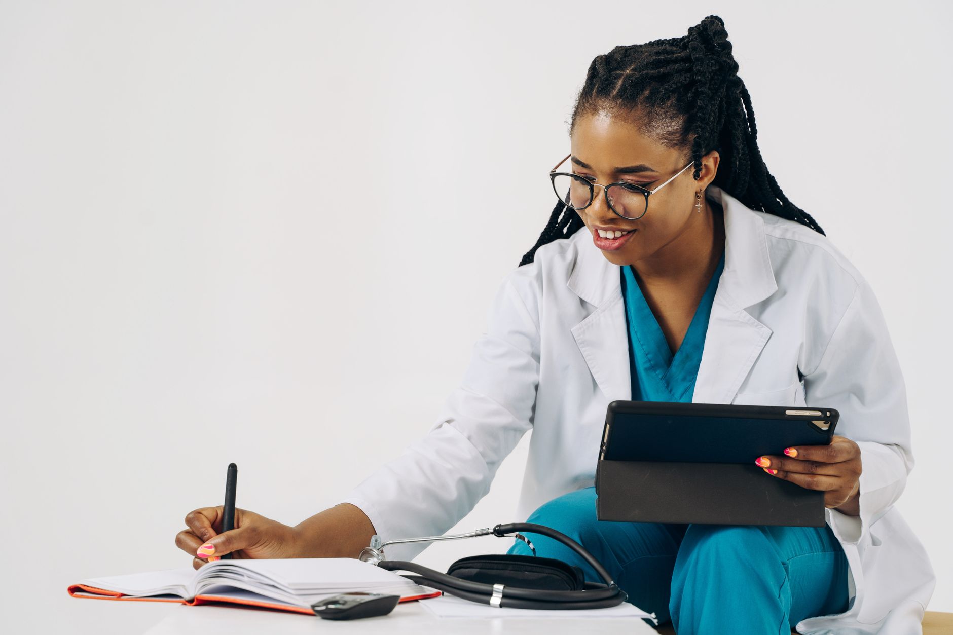 Smiling female doctor in scrubs using tablet and taking notes in a healthcare setting. (Photo by Tessy Agbonome on Pexels)