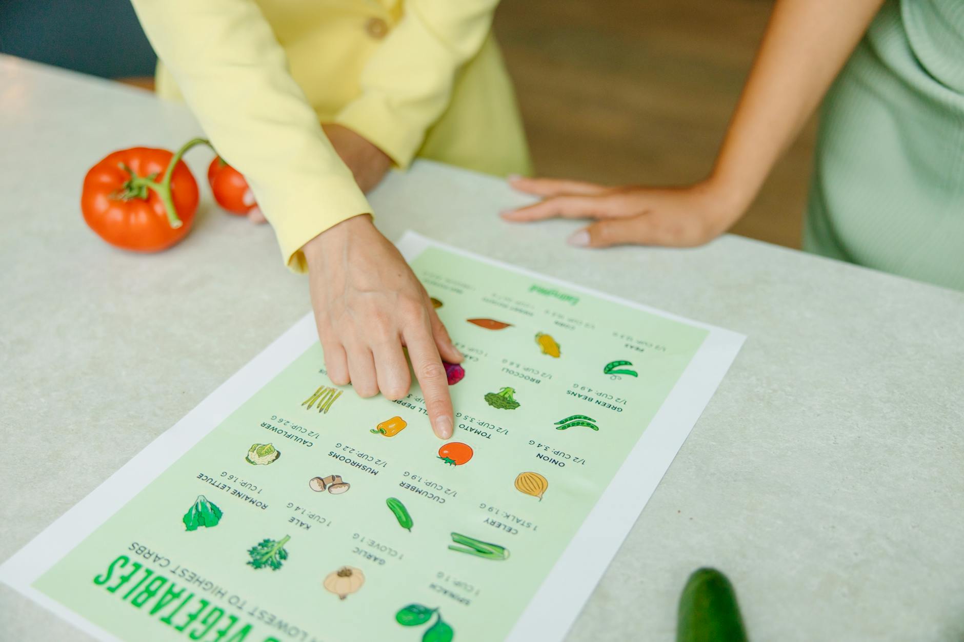 Close-up of hands pointing to a vegetable nutrition chart with fresh tomatoes on the table. (Photo by Yaroslav Shuraev on Pexels)